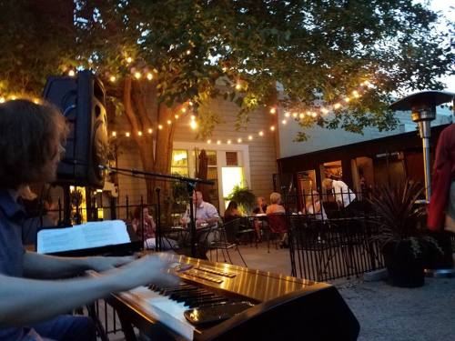 A musician plays piano outdoors at a lively restaurant, with string lights and diners enjoying the evening ambiance.