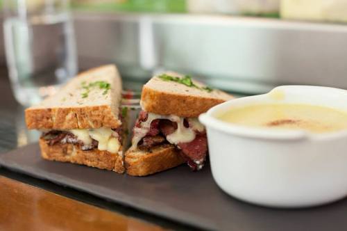 A close-up of a sandwich with layers of meat and cheese, served with a small bowl of creamy soup on a black plate.