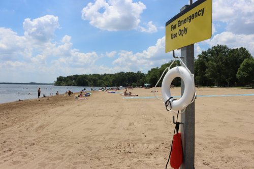 A sandy beach with people relaxing, a lifebuoy on a post labeled "For Emergency Use Only" in the foreground.