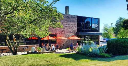 A sunny outdoor café scene with orange umbrellas, people sitting at tables, and a modern brick building in the background.