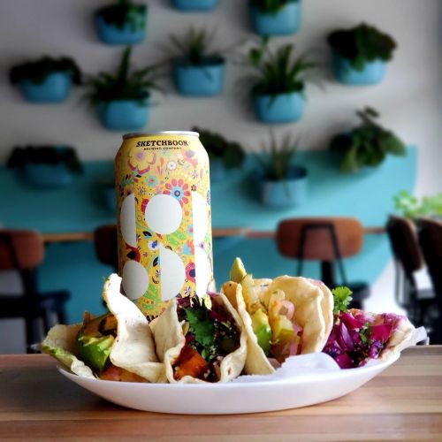A plate of colorful tacos with various toppings, next to a decorative can of beer, set against a vibrant background.