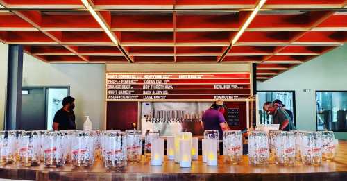 A bar with a wooden counter, several beer taps, and staff preparing drinks, with empty glasses lined up in front.