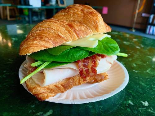 A croissant sandwich filled with turkey, bacon, spinach, and cheese, served on a paper plate.