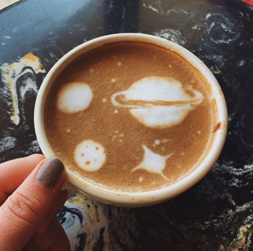 A hand holds a cup of coffee with latte art depicting planets and stars on the surface.