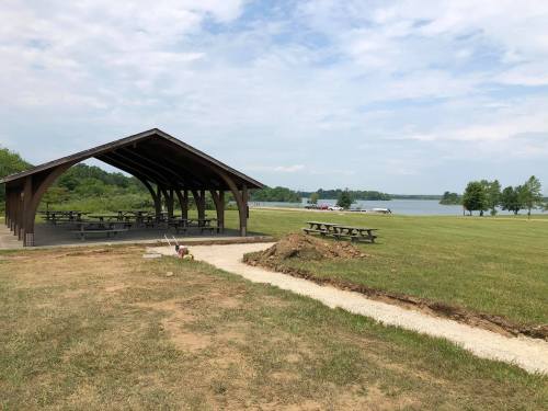 A picnic shelter near a lake, with picnic tables and a grassy area, under a partly cloudy sky.