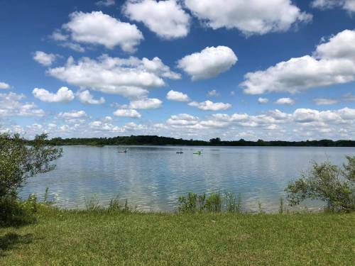 A serene lake scene with kayakers, surrounded by green grass and fluffy clouds in a blue sky.