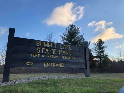 Sign for Summit Lake State Park with "Entrance" and "Dept. of Natural Resources" under a blue sky and scattered clouds.