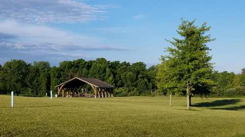 A wooden pavilion in a grassy field, surrounded by trees under a clear blue sky.