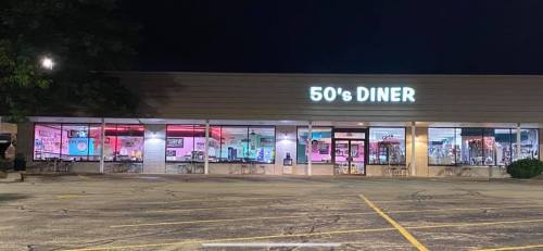 A brightly lit 50's diner at night, featuring large windows and retro decor, with a parking lot in front.