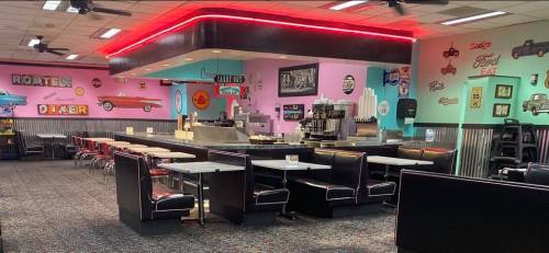 A retro diner interior with pink and teal walls, black booths, a counter, and vintage car decor.