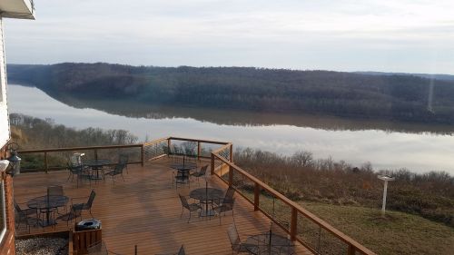A wooden deck with tables overlooks a calm river surrounded by hills and trees under a cloudy sky.