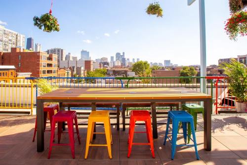 Colorful stools surround a large table on a rooftop with a city skyline in the background and hanging plants.