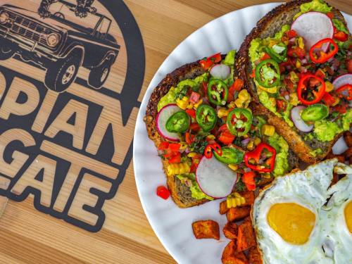 A plate of avocado toast topped with vegetables, alongside a fried egg and sweet potato cubes, with a logo in the background.