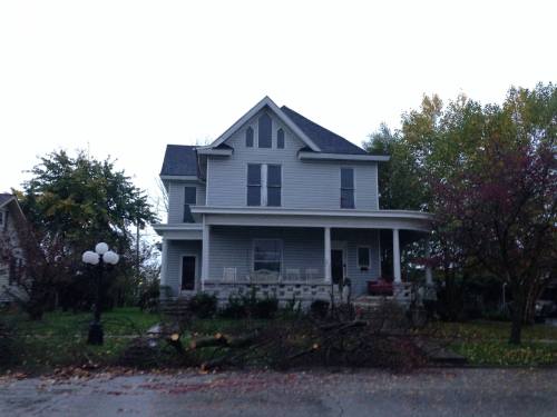 A gray two-story house with a porch, surrounded by fallen branches and trees, under a cloudy sky.