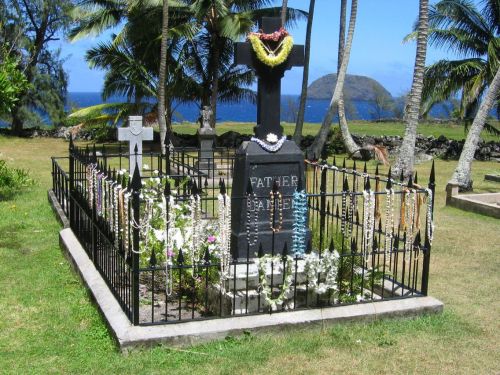A grave marked by a black cross, adorned with leis and flowers, surrounded by a wrought iron fence and palm trees.