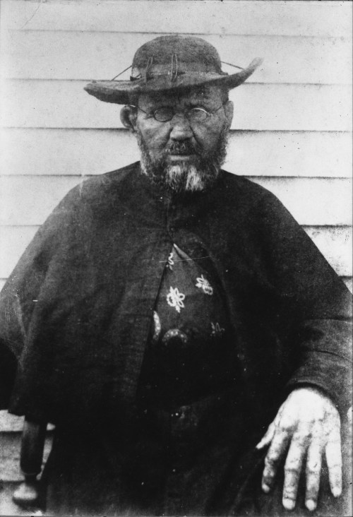 A bearded man in a hat and dark clothing sits, looking directly at the camera with a serious expression.