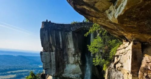 A scenic view of a cliff with a waterfall, surrounded by lush greenery and distant mountains under a clear blue sky.