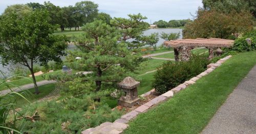A serene park scene featuring a lake, lush greenery, a stone pathway, and a decorative archway.
