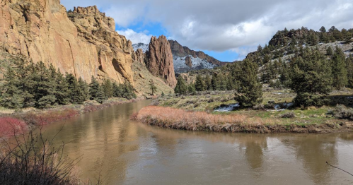 A winding river flows through a rocky canyon, surrounded by trees and snow-capped mountains under a partly cloudy sky.
