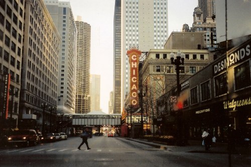 A bustling Chicago street scene with tall buildings, a vintage "Chicago" sign, and a pedestrian crossing the road.