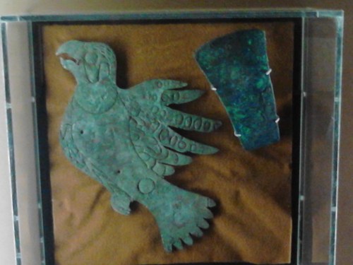 Bronze artifacts displayed in a glass case: a bird figure and a rectangular piece, both with intricate designs.