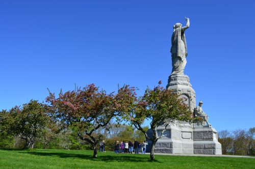 A tall statue on a grassy hill, with people gathered nearby and blooming trees in the foreground under a clear blue sky.