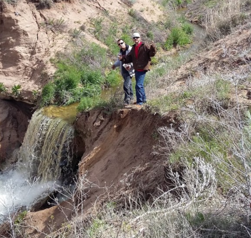 Two people stand near a small waterfall in a dry, grassy landscape with shrubs and a creek.