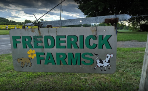 Sign for Frederick Farms featuring a flower, a cow, and a calf, set against a cloudy sky.