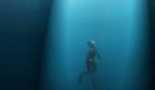 A diver swims underwater, illuminated by beams of light filtering through the surface above.