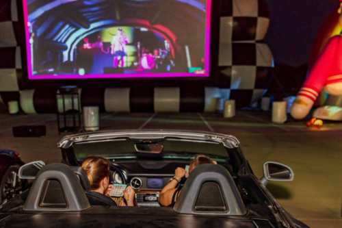 Two people sit in a convertible car, watching a large outdoor screen at a nighttime event. Colorful inflatable decorations nearby.