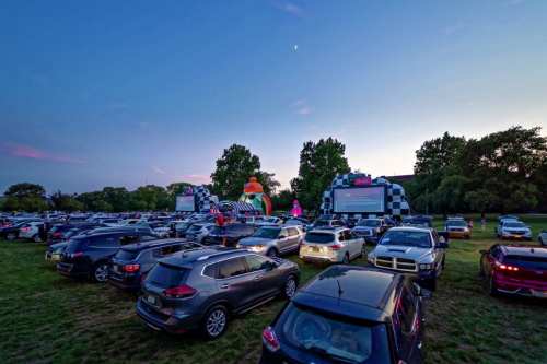 A drive-in movie scene at dusk, with cars parked and inflatable decorations in the background.