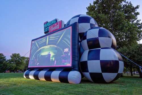 A large inflatable screen with a checkered pattern set up outdoors, displaying a live event in a park at dusk.
