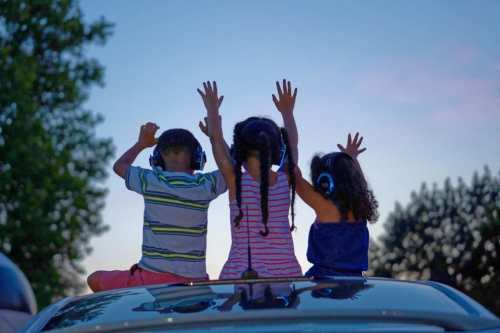 Three children with headphones sit on a car roof, raising their hands against a twilight sky.