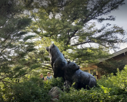 Two people stand near a large bear sculpture surrounded by greenery and trees.