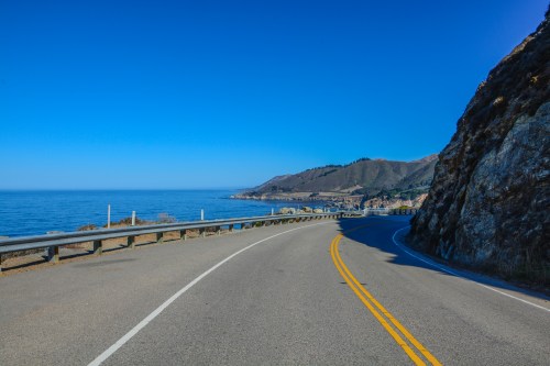 A winding coastal road curves along the ocean, with clear blue skies and rocky hills in the background.