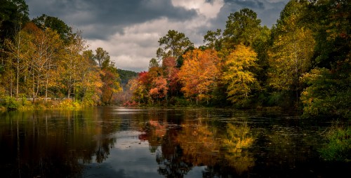 A serene river scene with vibrant autumn foliage reflecting on the water under a cloudy sky.