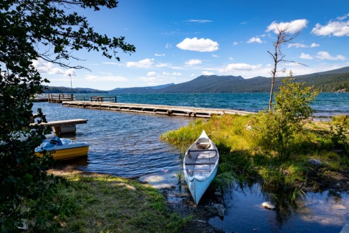 A serene lake scene with a canoe by the shore, a wooden dock, and mountains in the background under a blue sky.