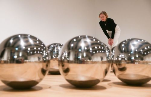 A woman in a black top leans over shiny metallic spheres in an art gallery, with reflections and lights in the background.