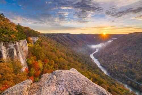 A scenic view of a river winding through a colorful forest at sunrise, with cliffs in the foreground and mist in the valley.