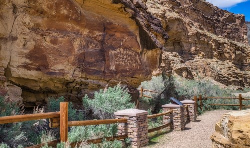 A scenic pathway with stone pillars and a wooden fence, surrounded by rocky cliffs and greenery.