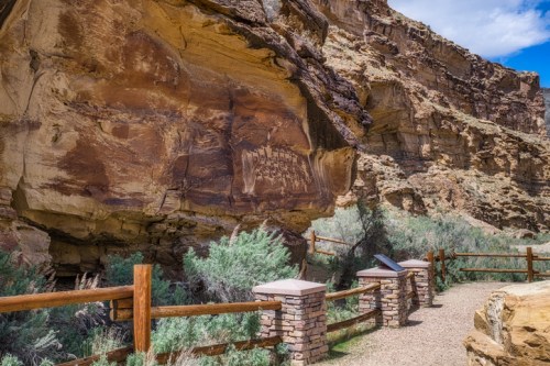 Rocky canyon landscape with ancient petroglyphs on a cliff, surrounded by shrubs and a wooden fence. Clear blue sky above.