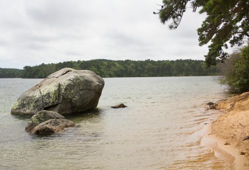 A large rock sits near the water's edge, with a sandy shore and trees in the background under a cloudy sky.