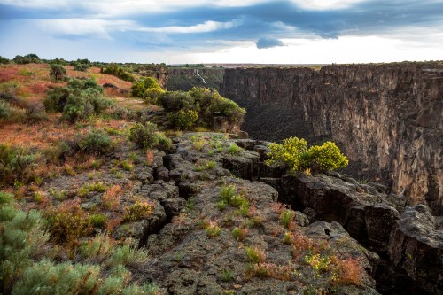 A rocky cliffside overlooks a deep canyon, surrounded by lush greenery and dramatic clouds in the sky.