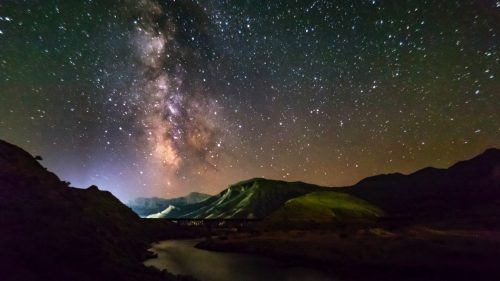 A stunning night sky filled with stars and the Milky Way over a serene landscape with mountains and a river.