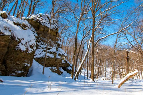 Snow-covered landscape featuring rocky outcrops and bare trees under a clear blue sky.