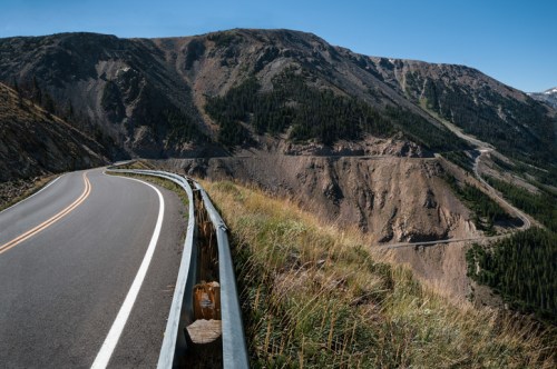 A winding road curves along a mountainside, with steep slopes and greenery in the background under a clear blue sky.