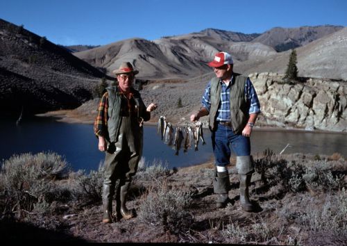 Two men in fishing gear hold a string of caught fish by a lake, with mountains in the background under a clear blue sky.