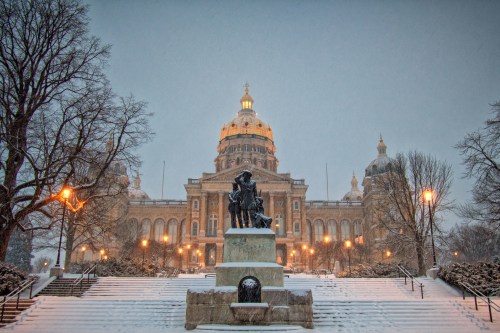 Snow falls on a grand building with a dome, framed by trees and a statue in the foreground, creating a serene winter scene.