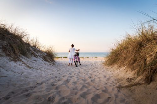 A person in a white dress stands with a bicycle at the beach, framed by tall grass and sandy dunes.
