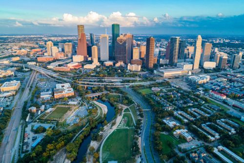Aerial view of a vibrant city skyline with skyscrapers, green parks, and winding roads under a blue sky.